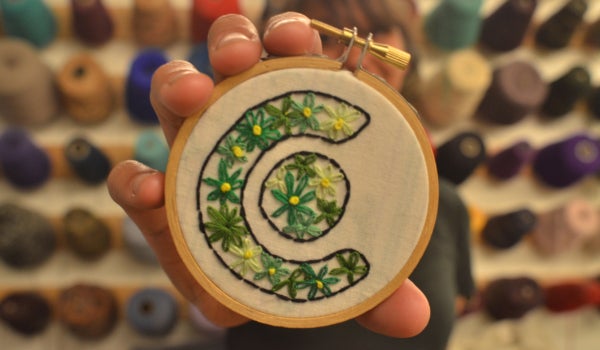 Someone holds up the embroidery they finished of the Craft Center logo composed of flowers.